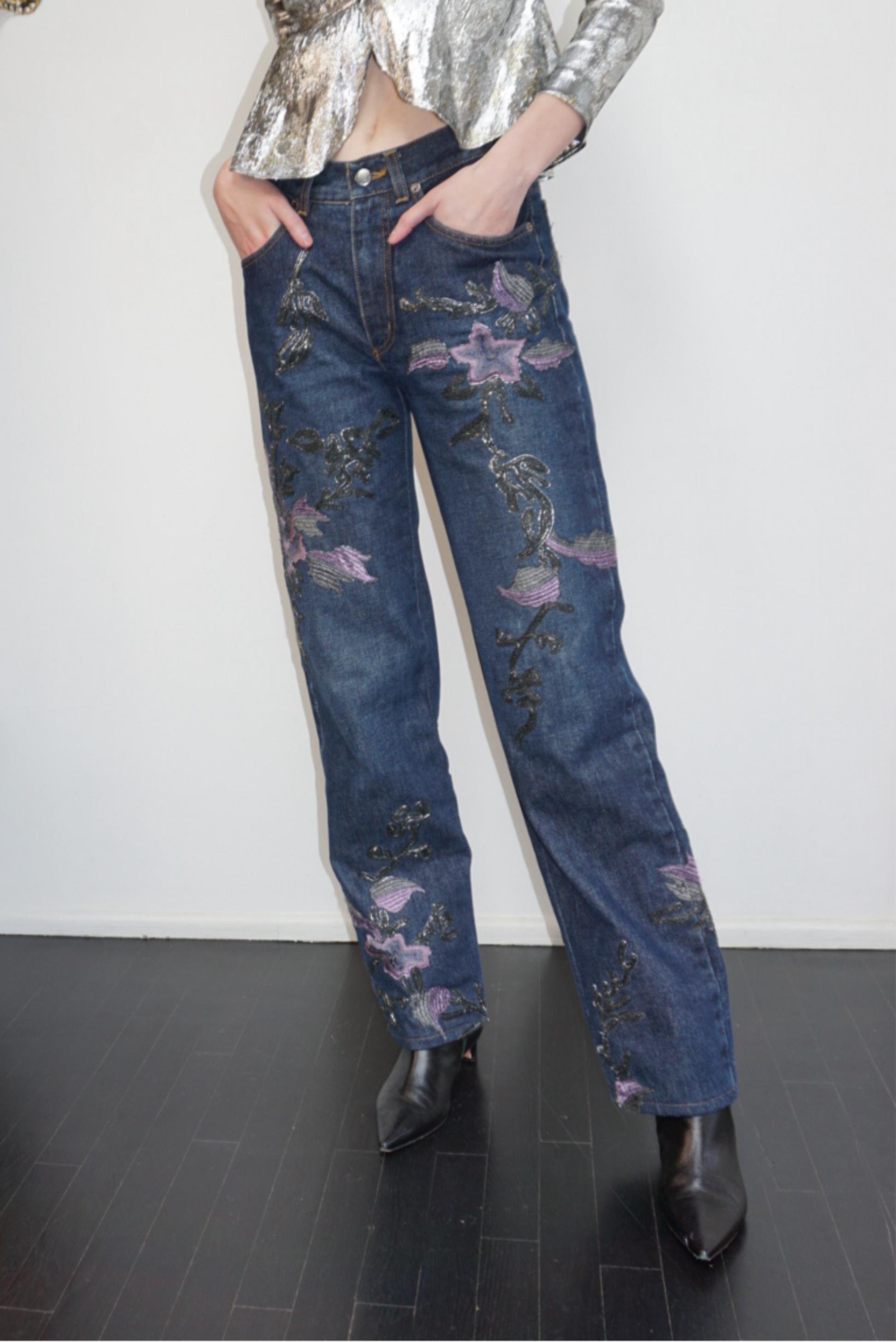 Model sitting wearing denim jeans with metallic soutache embroidery and purple flowers, a sequin top and black boots.