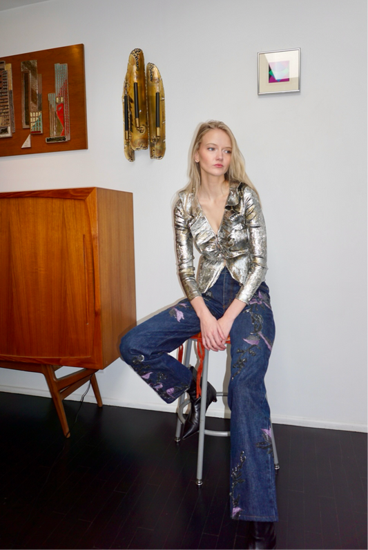 Model wearing denim jeans with metallic soutache embroidery and purple flowers, a sequin top and black boots.