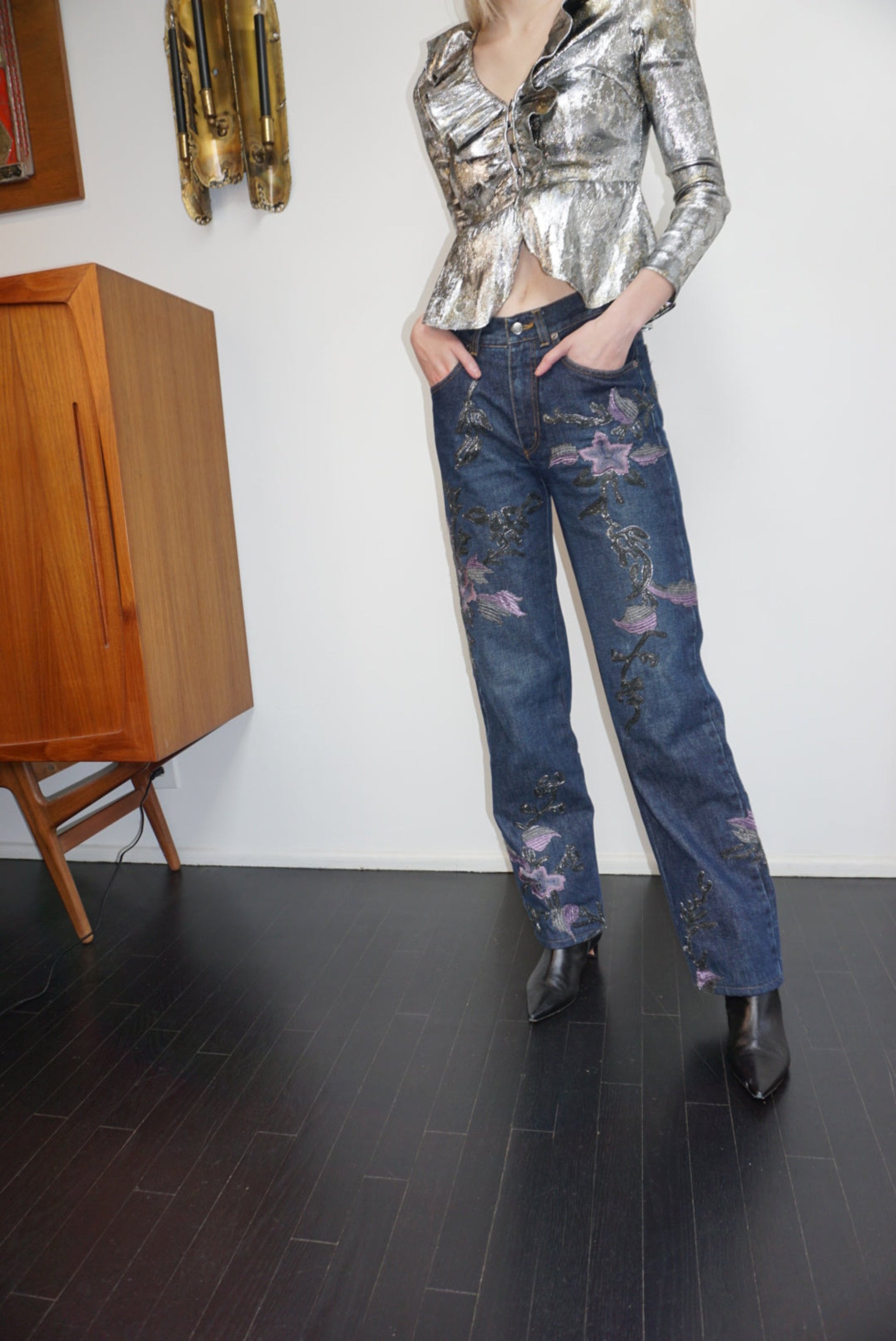 Model sitting wearing denim jeans with metallic soutache embroidery and purple flowers, a sequin top and black boots.