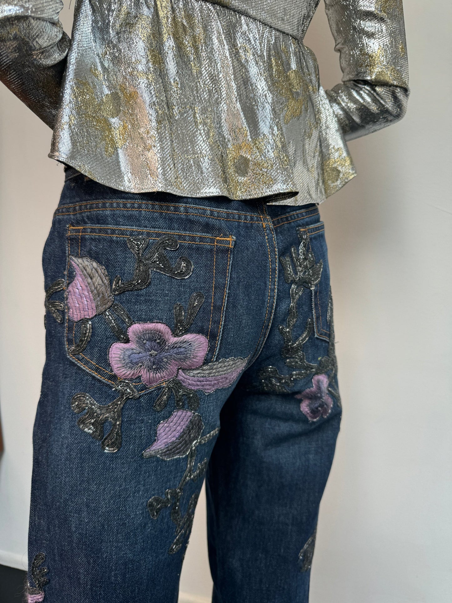 The back of a model wearing a silver shirt and denim jeans with metallic soutache embroidery and purple flowers.