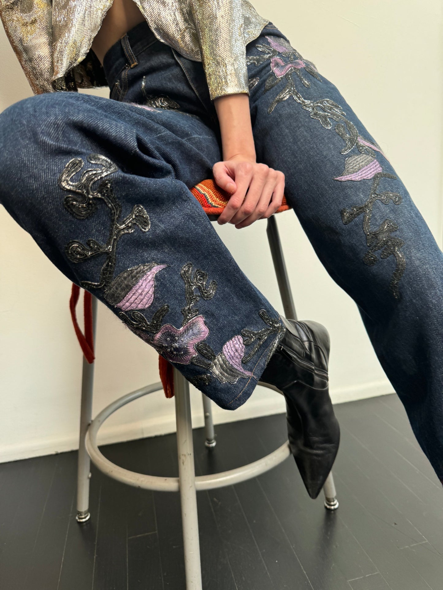 Model sitting wearing denim jeans with metallic soutache embroidery and purple flowers, a sequin top and black boots.