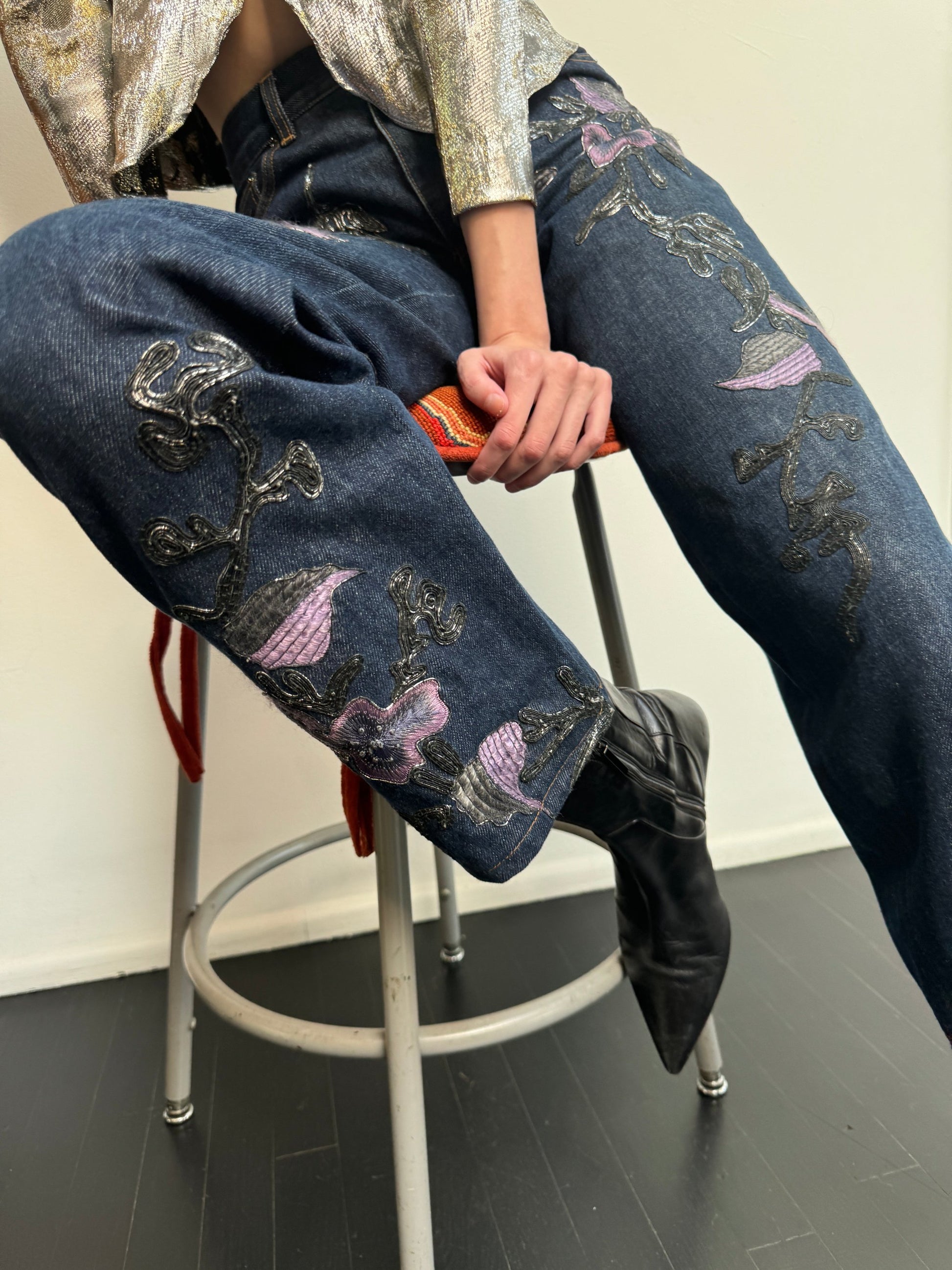 Model sitting wearing denim jeans with metallic soutache embroidery and purple flowers, a sequin top and black boots.