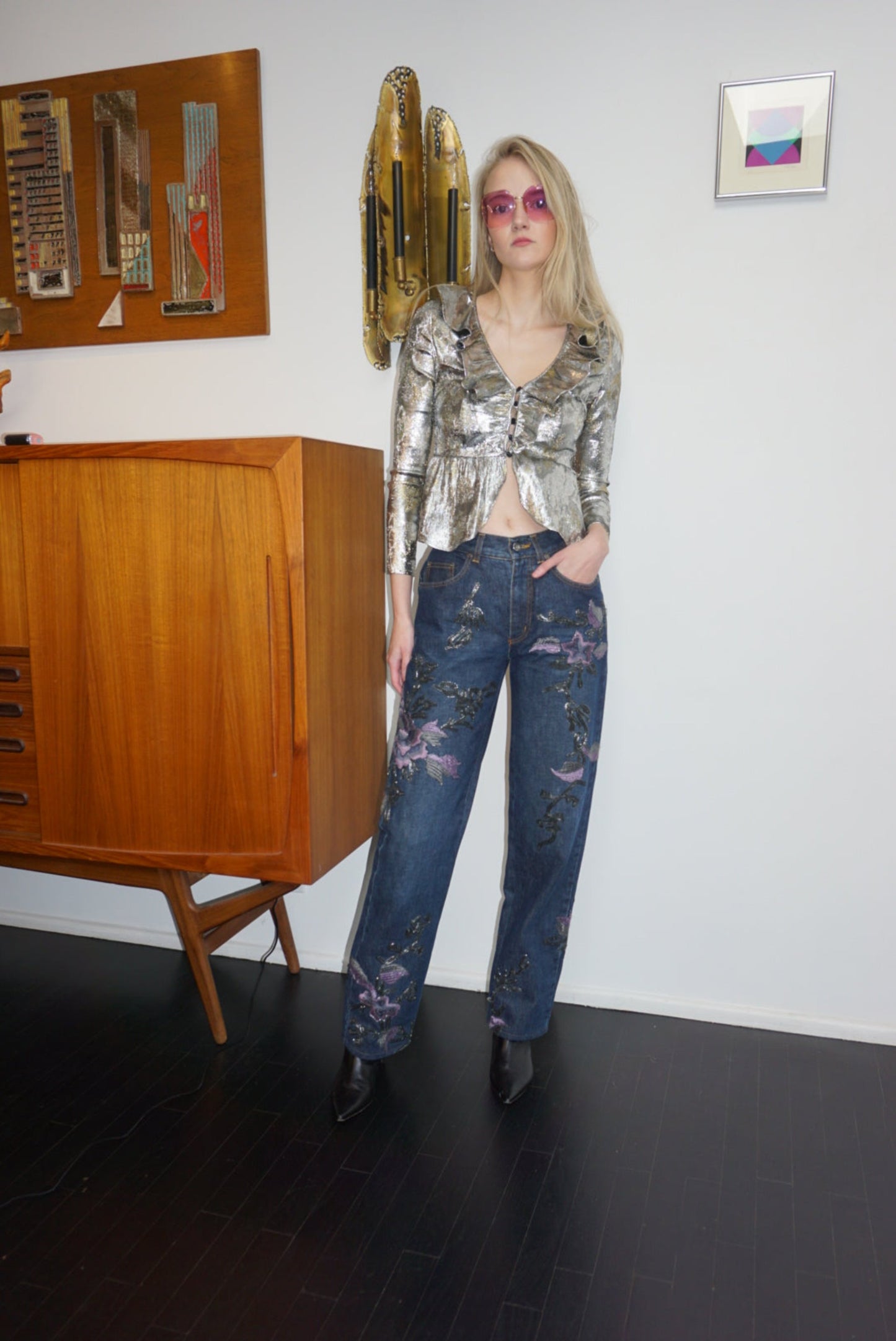 A model wears a silver sequin top, denim jeans with metallic soutache embroidery and purple flowers.